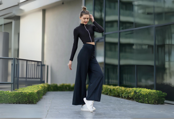 Frau posiert in schwarzem Outfit und weißen Turnschuhen auf dem Gehweg eines modernen Gebäudes mit viel Grün.